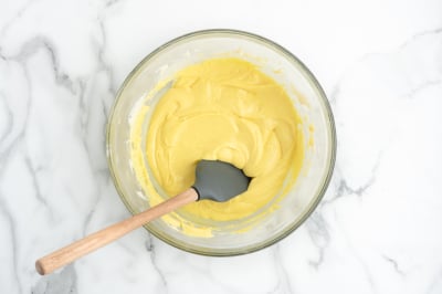 A glass bowl filled with creamy yellow batter for a delicious Lemon Fudge Recipe sits on a white marble surface, with a black silicone spatula with a wooden handle resting inside.