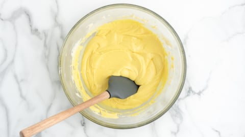 A glass bowl filled with creamy yellow batter for a delicious Lemon Fudge Recipe sits on a white marble surface, with a black silicone spatula with a wooden handle resting inside.