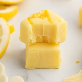 Two creamy yellow fudge squares are stacked, with the top piece showing bite marks. Lemon slices and white chocolate chips blur in the background, hinting at a zesty Lemon Fudge Recipe.