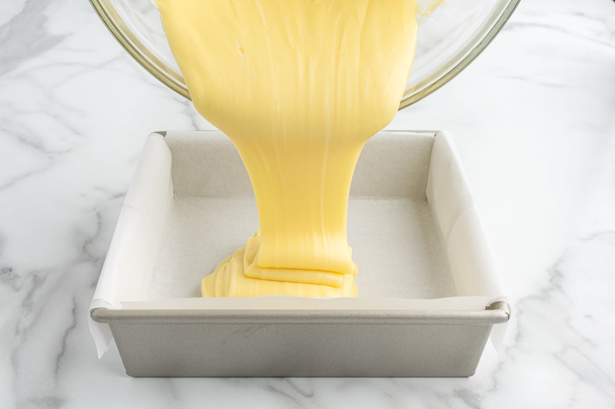 Yellow cake batter, perfect for a Lemon Fudge Recipe, is being poured from a glass bowl into a square metal baking pan lined with parchment paper, all set on a white marble countertop.