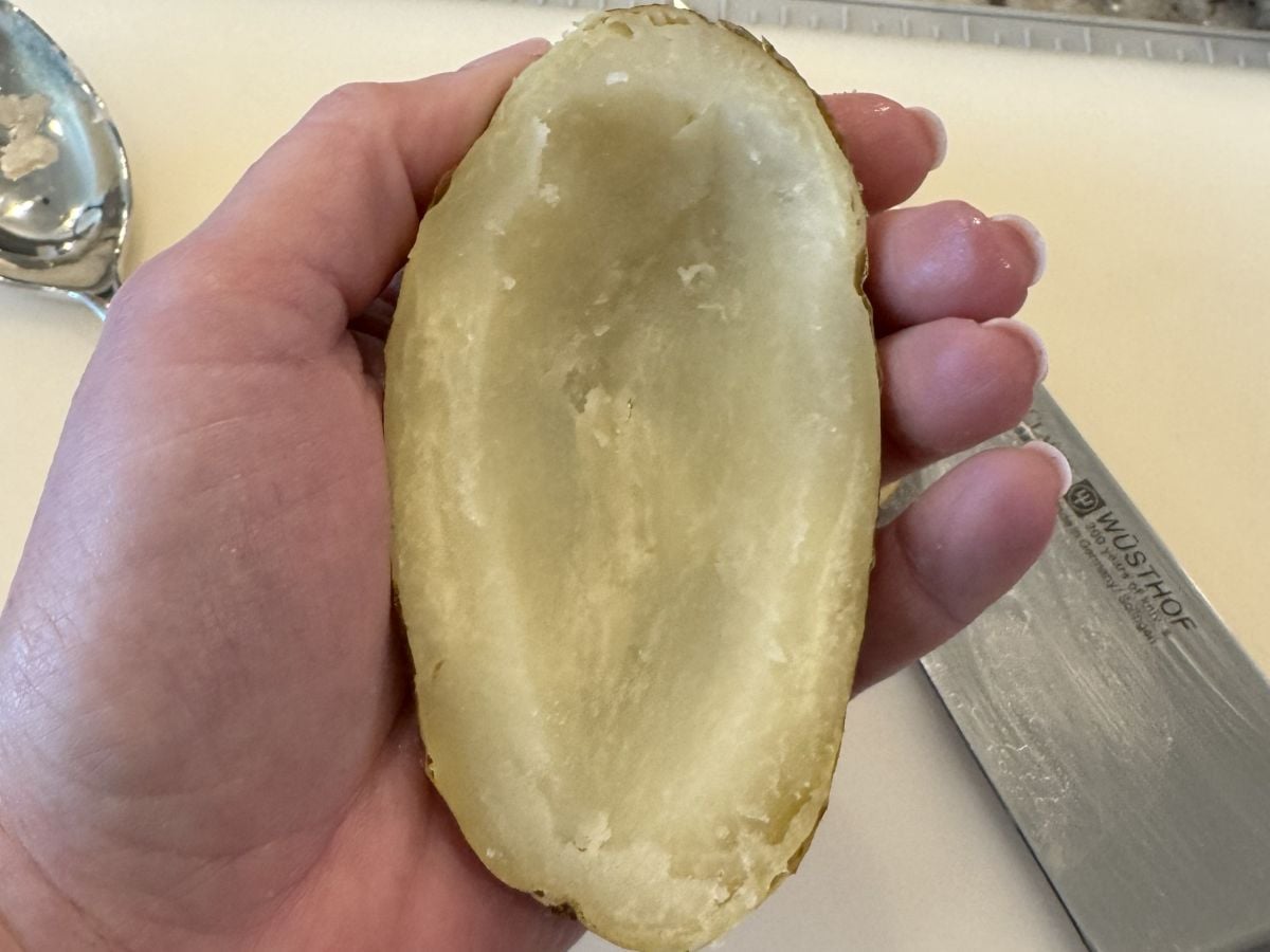 A hand holds half of a baked potato skin with the inside scooped out, leaving just the skin and a thin layer of potato flesh. A metal knife and spoon rest on a white surface in the background.