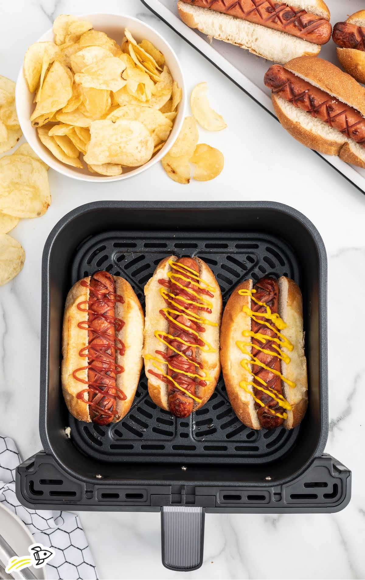 Hot Dogs topped with ketchup and mustard in an air fryer next to a bowl of chips