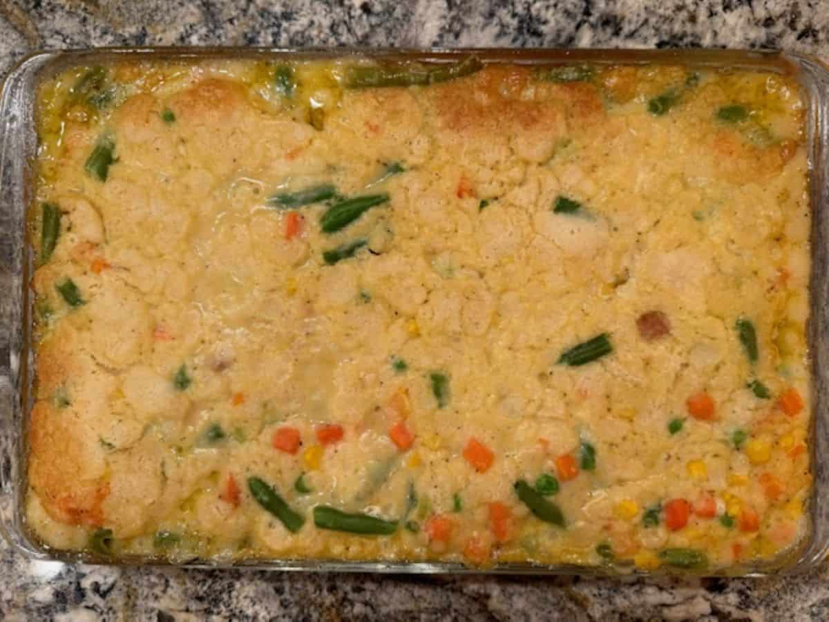 A Chicken Cobbler baked casserole with a golden biscuit-like topping, mixed with visible vegetables like green beans, carrots, and corn, sits on a granite countertop.