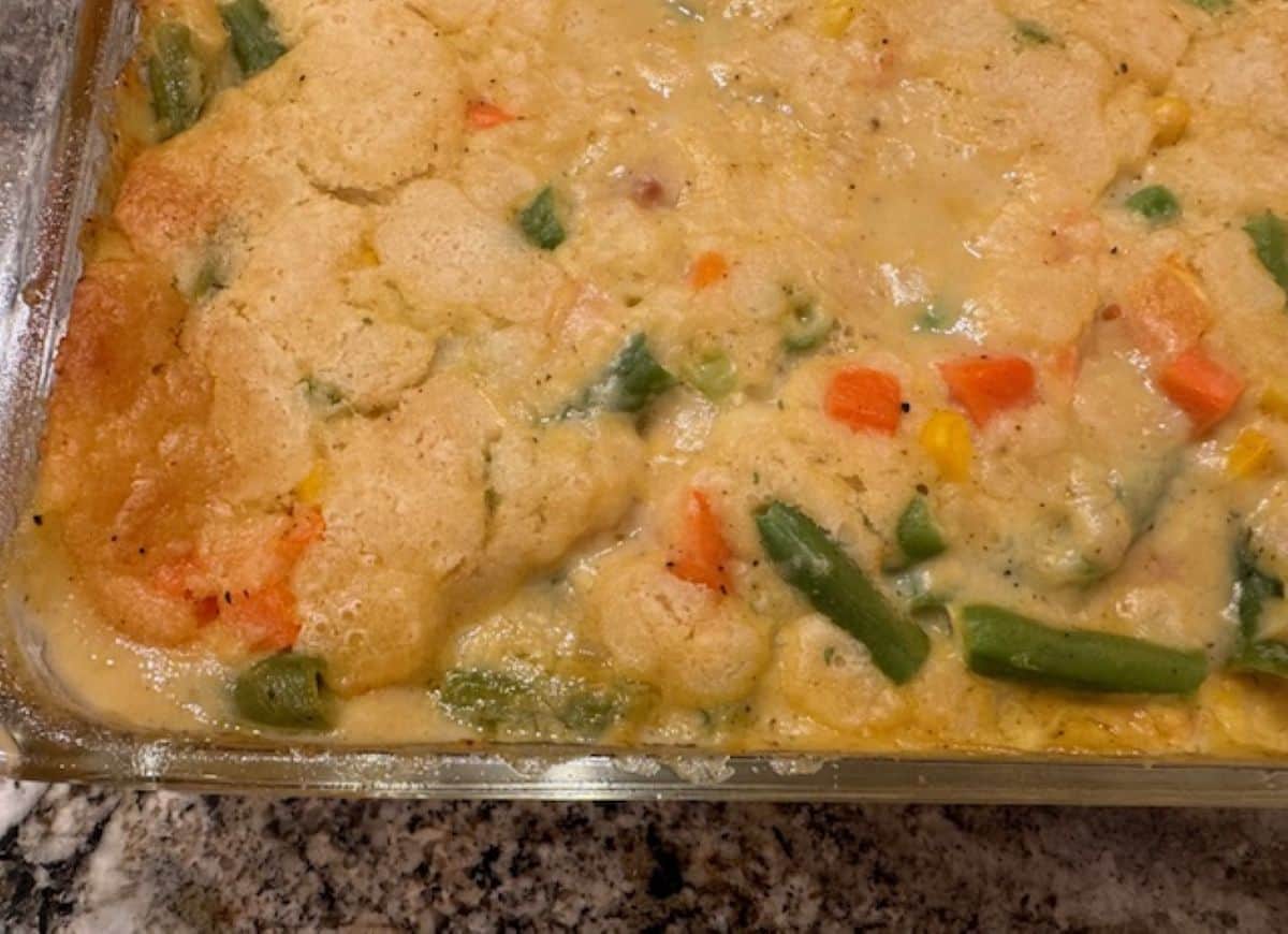 A glass baking dish filled with a creamy vegetable casserole, reminiscent of a comforting Chicken Cobbler, featuring a golden biscuit topping and visible pieces of carrots, green beans, and corn.