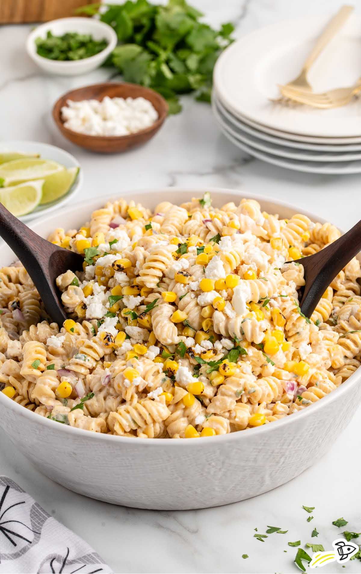 Side angle of a bowl of Mexican street corn pasta salad with wooden serving spoons, surrounded by lime wedges, cilantro, and stacked plates.