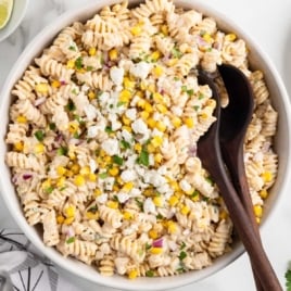 Top-down image of a finished bowl of Mexican street corn pasta salad with dark wooden serving utensils and a sprinkle of cotija cheese on top.