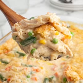A close-up of a wooden spoon holding a serving of creamy Chicken Cobbler casserole with green beans, corn, peas, carrots, and a golden, crispy topping.