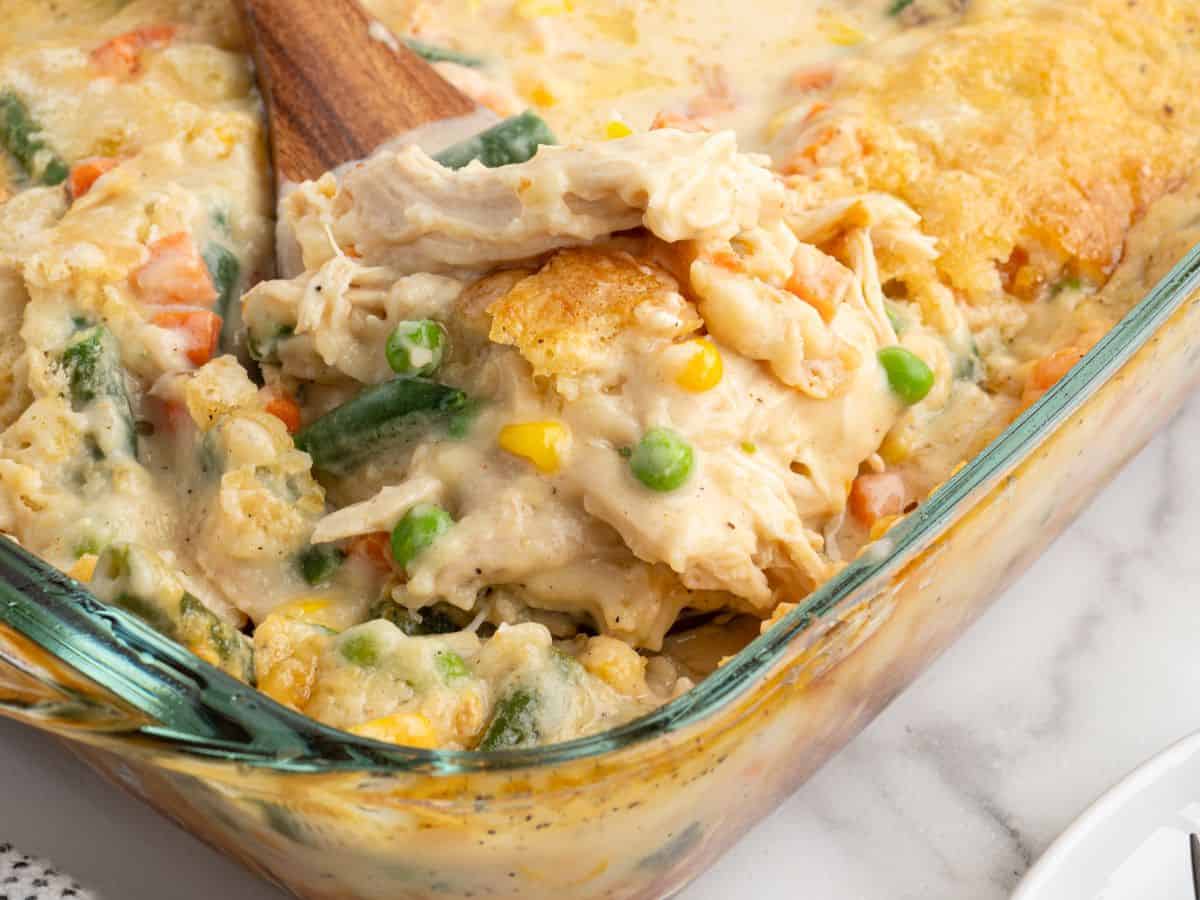 A close-up of a baking dish filled with creamy Chicken Cobbler casserole, mixed vegetables, and a golden biscuit topping. A wooden spoon is lifting a serving from the dish.