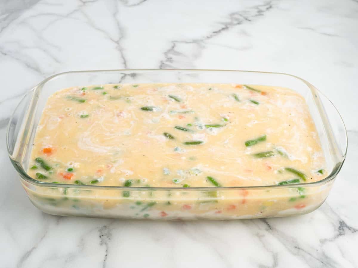 A glass baking dish filled with an uncooked Chicken Cobbler casserole mixture containing visible chopped vegetables sits on a marble countertop.