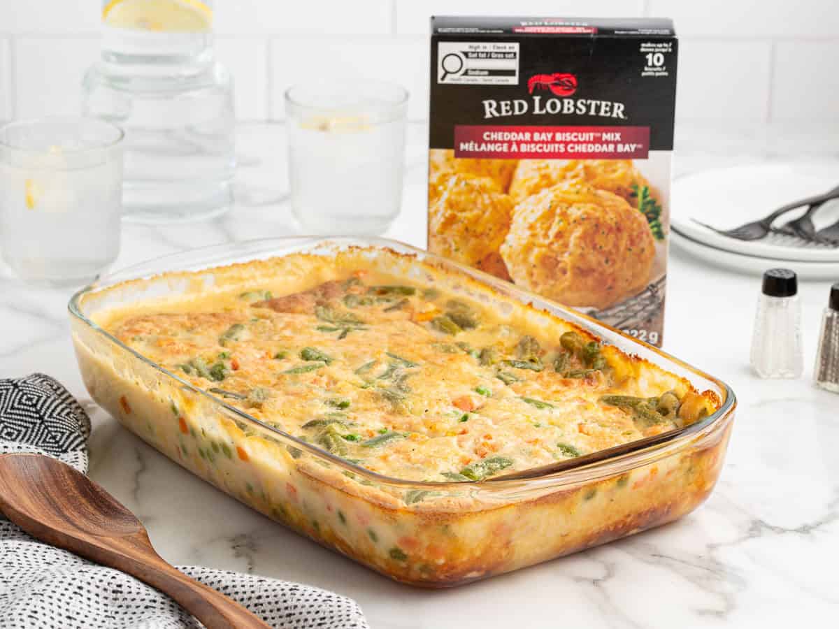A Chicken Cobbler baked casserole in a glass dish sits on a marble counter near a Red Lobster Cheddar Bay Biscuit Mix box, with utensils, water glasses, and a napkin nearby.