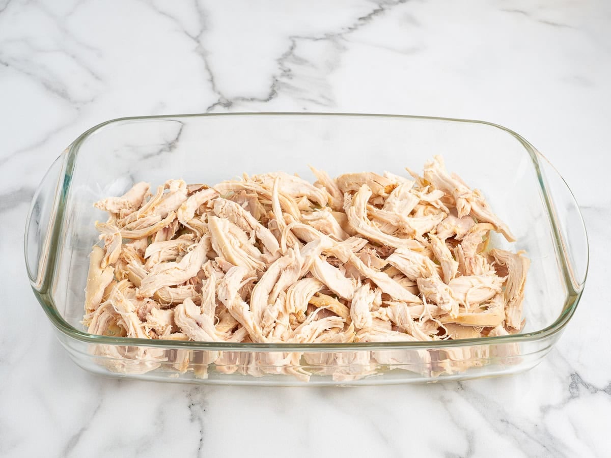A glass baking dish filled with shredded cooked chicken, reminiscent of a hearty Chicken Cobbler, set on a white marble countertop.