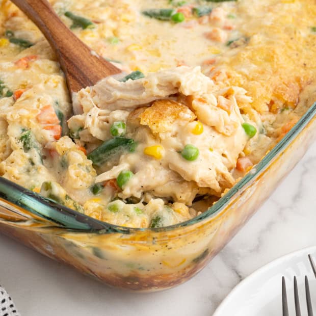A glass baking dish filled with creamy Chicken Cobbler pot pie casserole, topped with a golden crust, with a wooden spoon lifting a serving. Green beans, peas, carrots, and corn are visible. Plates and forks are nearby.