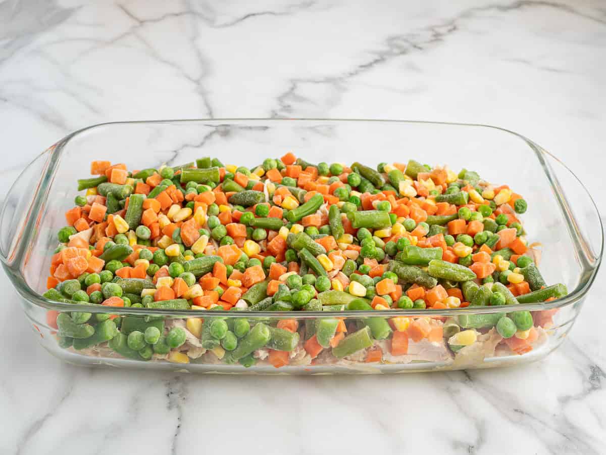 A glass baking dish filled with a colorful mix of frozen vegetables, including green beans, peas, corn, and diced carrots, sits on a white marble countertop—perfect as the base for a hearty Chicken Cobbler.