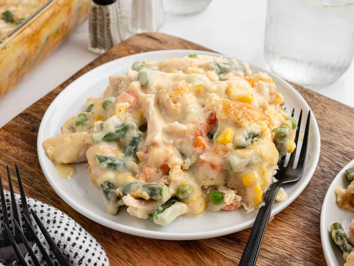 A serving of creamy Chicken Cobbler casserole with mixed vegetables on a white plate, placed on a wooden board with a black fork. A glass of water and casserole dish are in the background.