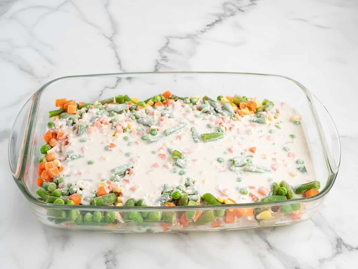 A glass baking dish filled with mixed frozen vegetables and a creamy white sauce, reminiscent of the comforting flavors of Chicken Cobbler, sits on a white marble countertop.