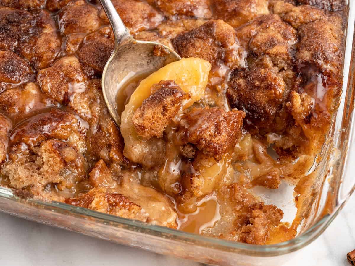 A close-up of a Snickerdoodle Cobbler in a glass baking dish, with a spoon scooping out a portion, revealing juicy peaches and a golden, cinnamon-topped biscuit crust.