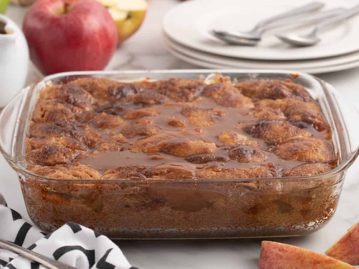 A glass baking dish filled with Snickerdoodle Cobbler, topped with a glossy brown caramel sauce, set on a table with apples, plates, and utensils nearby.