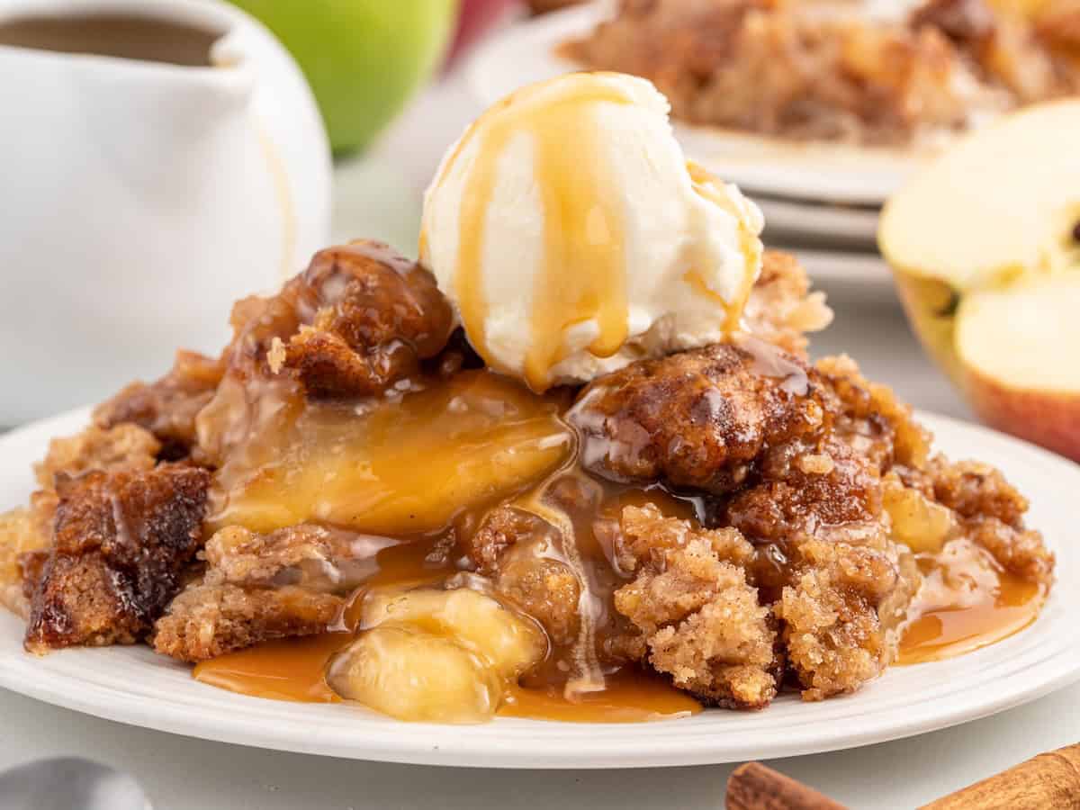 A plate of Snickerdoodle Cobbler crumble topped with a scoop of vanilla ice cream and drizzled with caramel sauce, with apple slices and a sauce pitcher in the background.
