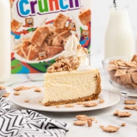 a slice of Cinnamon Toast Crunch Cheesecake topped with whipped topping and cinnamon toast crunch cereal crumbs on a plate