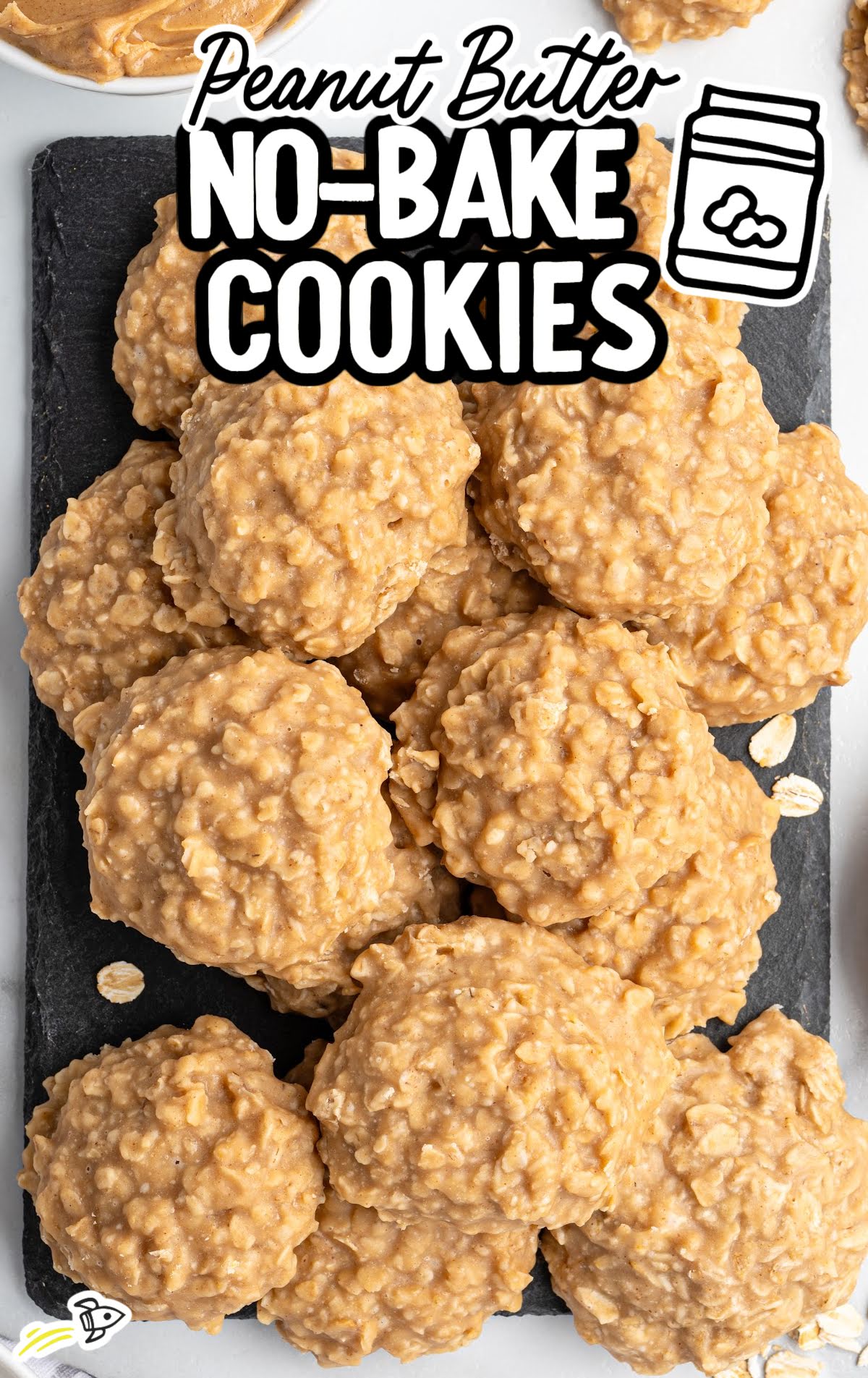 Close-up overhead image of a pile of no-bake peanut butter cookies on a black slate board, with bold text reading 'Peanut Butter No-Bake Cookies' and a jar icon.