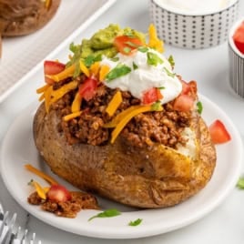 A baked potato inspired by Baked Potato Tacos, topped with chili, shredded cheddar cheese, diced tomatoes, guacamole, sour cream, and chopped cilantro, served on a white plate.