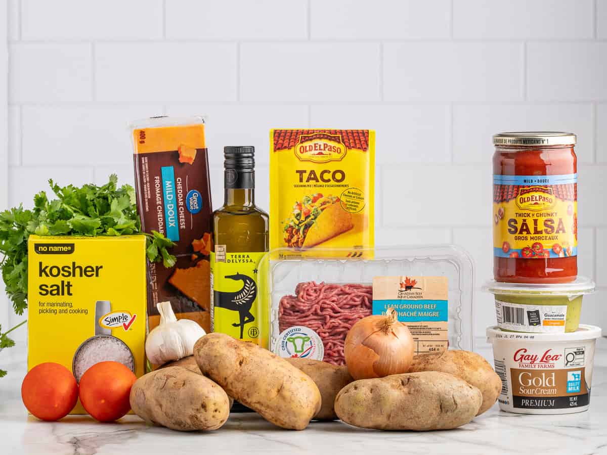 A variety of taco ingredients for Baked Potato Tacos on a counter, including ground beef, potatoes, onion, tomatoes, cilantro, salt, garlic, olive oil, taco shells, salsa, sour cream, and taco seasoning against a white tiled background.