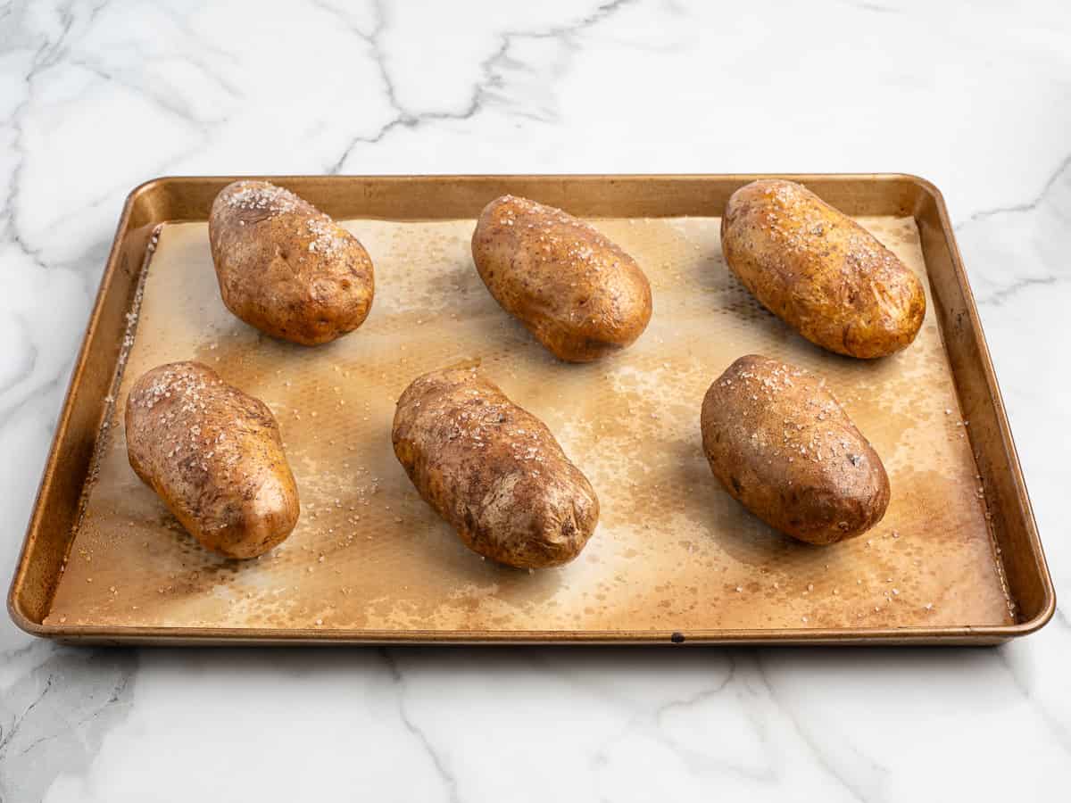 Six whole, unpeeled potatoes sprinkled with salt are arranged evenly on a baking sheet lined with parchment paper, ready to be transformed into delicious Baked Potato Tacos on a white marble surface.