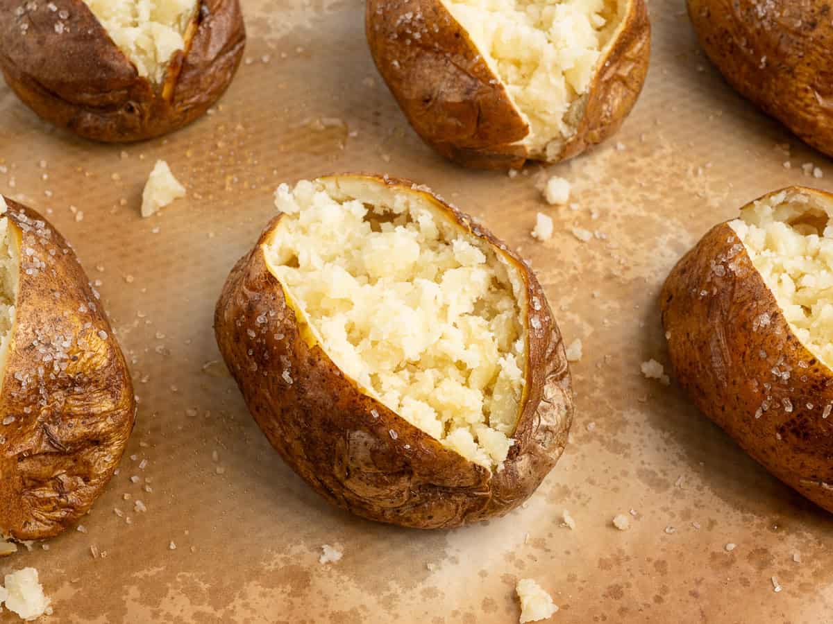 Several baked potatoes with crispy skins are split open and stuffed taco-style, inspired by baked potato tacos. They rest on brown parchment paper with scattered potato crumbs and coarse salt.