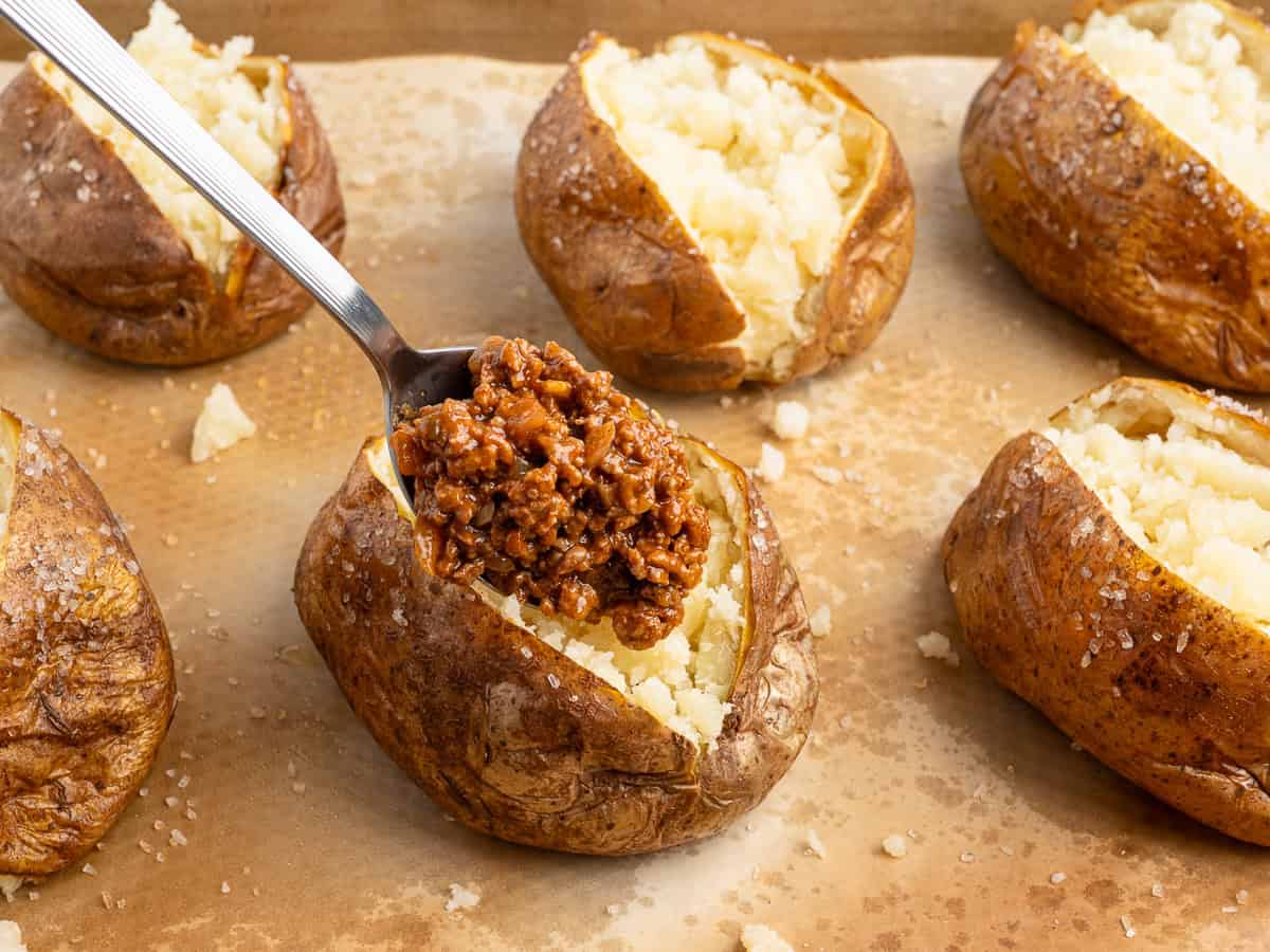 A spoonful of chili is being added to a baked potato with a split, fluffy inside, reminiscent of the heartiness found in Baked Potato Tacos. Several other salted baked potatoes rest on a parchment-lined baking sheet.