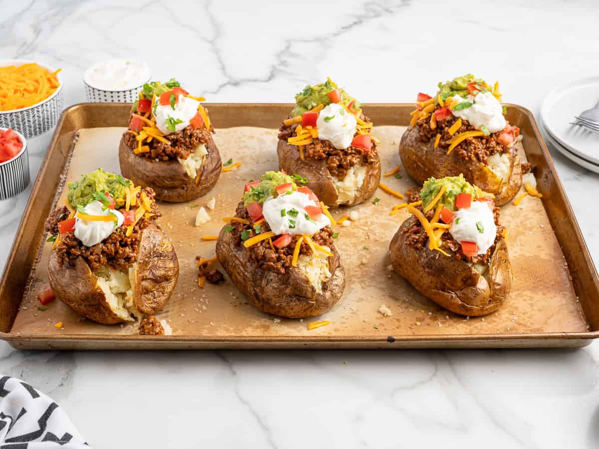 Six baked potatoes on a baking sheet, each split open and piled high like baked potato tacos with ground meat, shredded cheese, diced tomatoes, guacamole, sour cream, and chopped cilantro. Bowls of toppings are visible in the background.