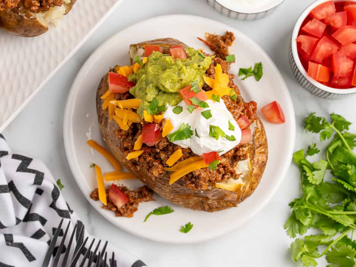 A baked potato taco topped with ground meat chili, shredded cheddar cheese, sour cream, guacamole, chopped tomatoes, and fresh cilantro, served on a white plate with a side of diced tomatoes and cilantro.