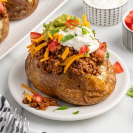 A baked potato taco twist: a baked potato topped with chili, shredded cheddar cheese, diced tomatoes, guacamole, sour cream, and chopped cilantro. Served on a white plate with bowls of sour cream and tomatoes in the background.