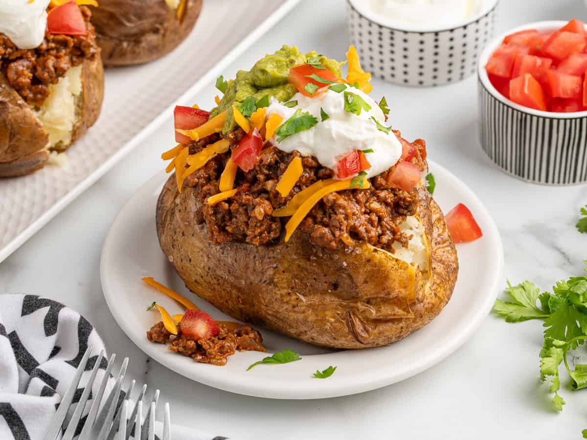 A baked potato taco twist: a baked potato topped with chili, shredded cheddar cheese, diced tomatoes, guacamole, sour cream, and chopped cilantro. Served on a white plate with bowls of sour cream and tomatoes in the background.