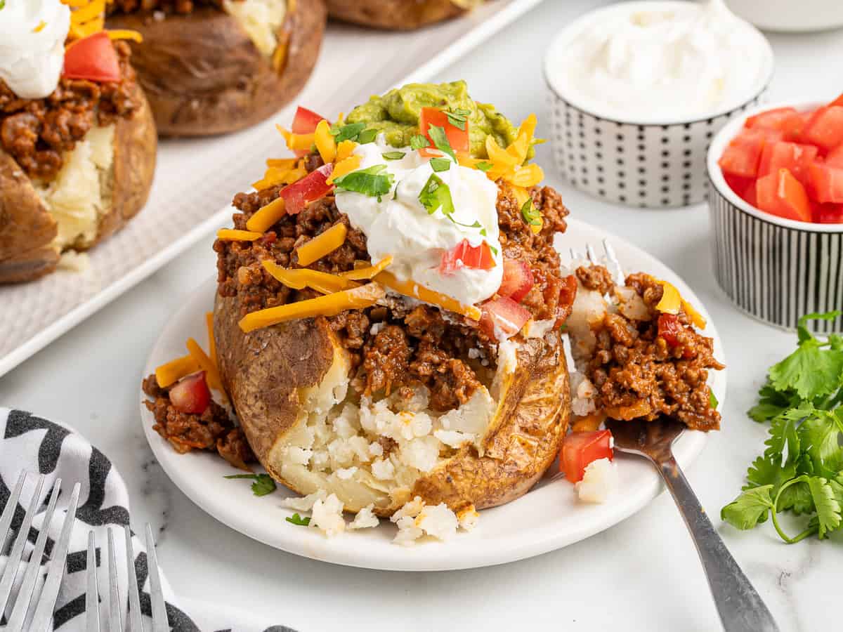 A baked potato split open and topped with chili, shredded cheese, diced tomatoes, guacamole, sour cream, and chopped cilantro sits on a white plate with a fork—like the perfect fusion of Baked Potato Tacos. Bowls of tomatoes and sour cream are in the background.