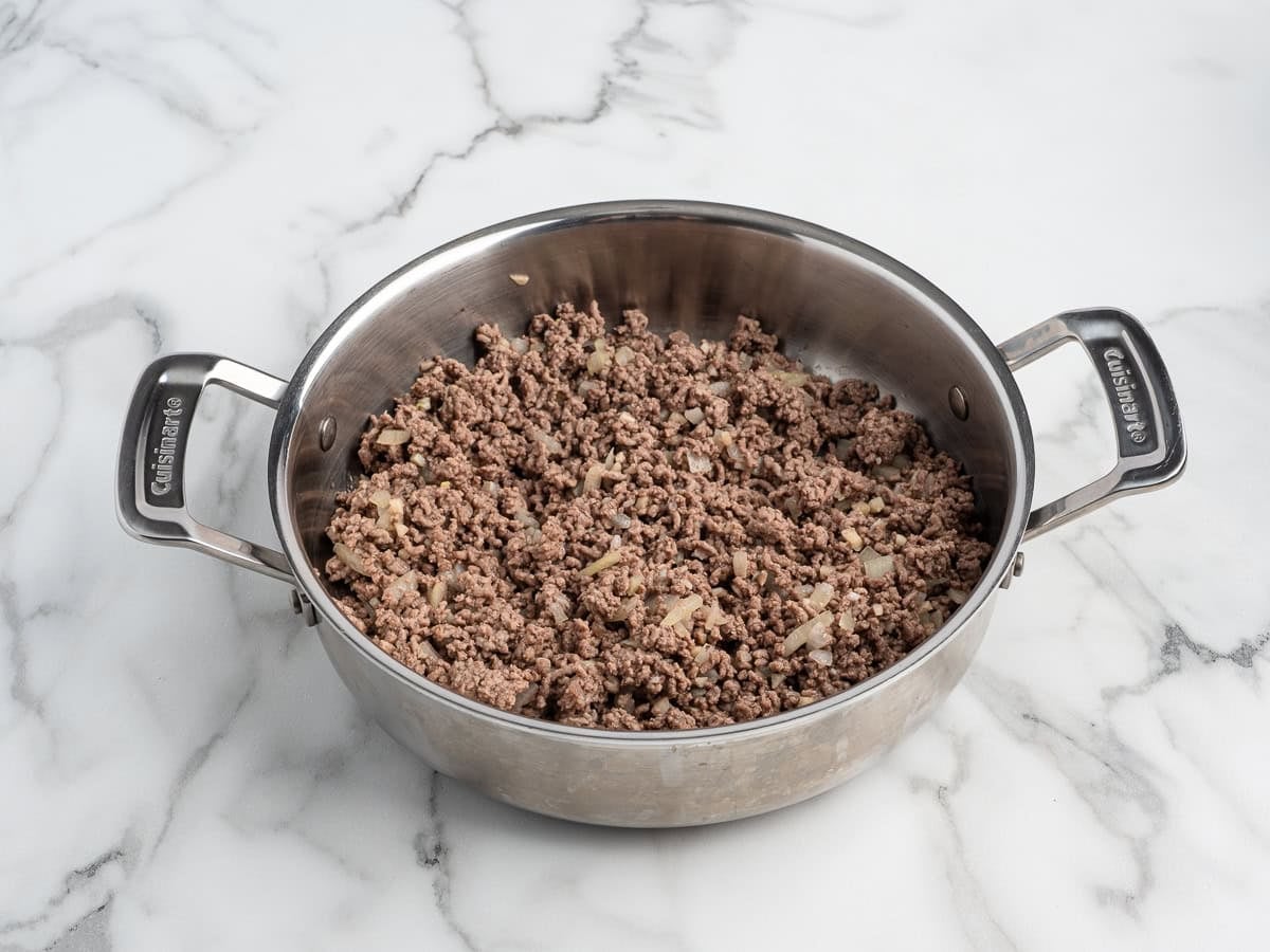 A stainless steel pan with cooked ground beef and chopped onions sits on a white marble countertop, ready to be used as a savory filling for Baked Potato Tacos.