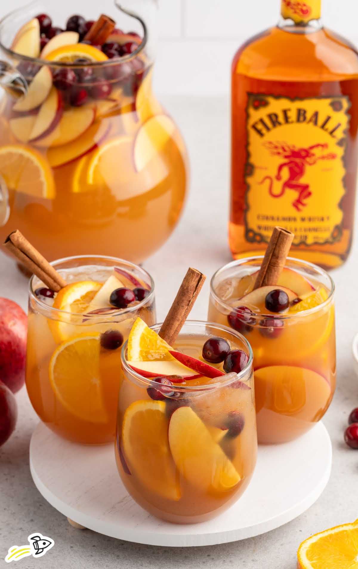 Three glasses of Fireball apple cider sangria with fruit slices and cinnamon sticks next to a large pitcher and bottle of Fireball.