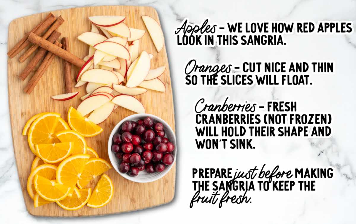 Cutting board with sliced red apples, orange slices, cinnamon sticks, and a bowl of cranberries prepared for Fireball sangria.