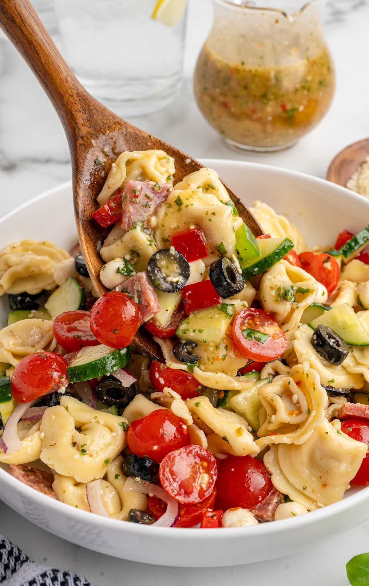 Wooden spoon lifting a serving of tortellini pasta salad featuring tomatoes, cucumbers, olives, and salami.