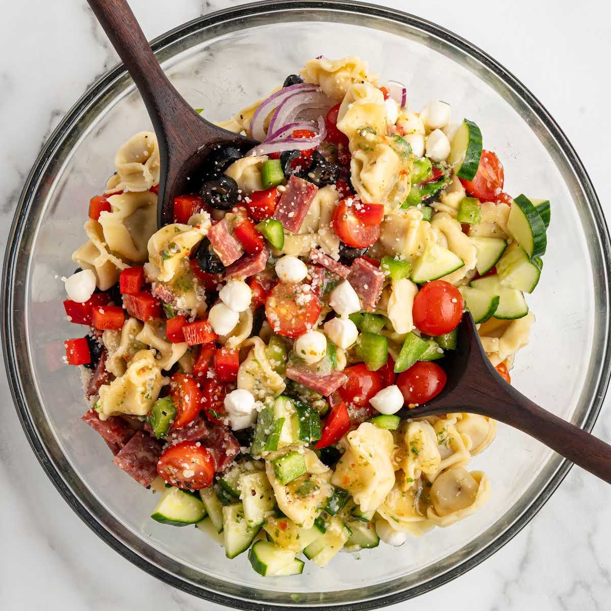 Overhead view of tortellini pasta salad in a glass bowl with vegetables, salami, mozzarella pearls, and Italian dressing.