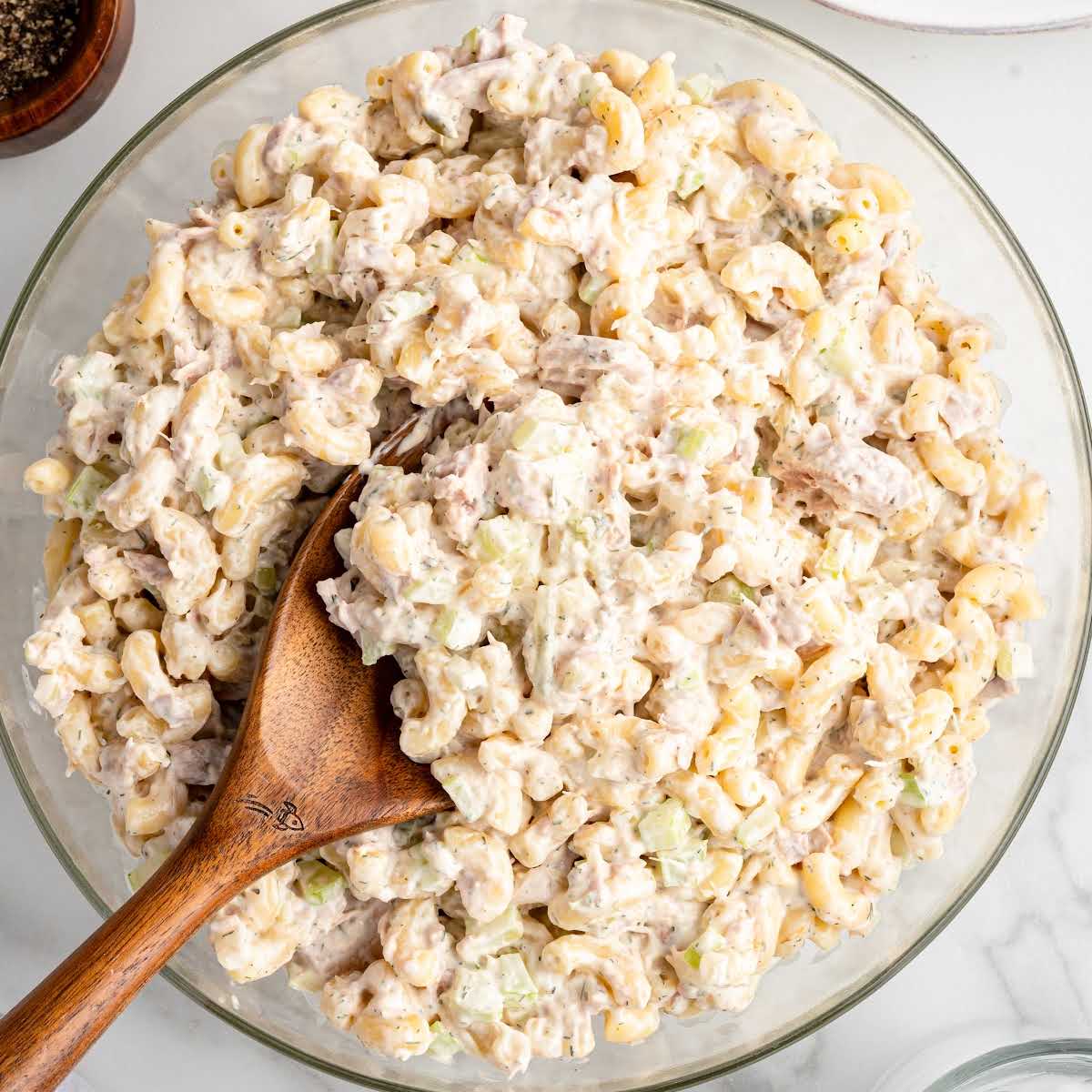 Overhead view of finished tuna macaroni salad in a glass bowl with a wooden spoon for serving