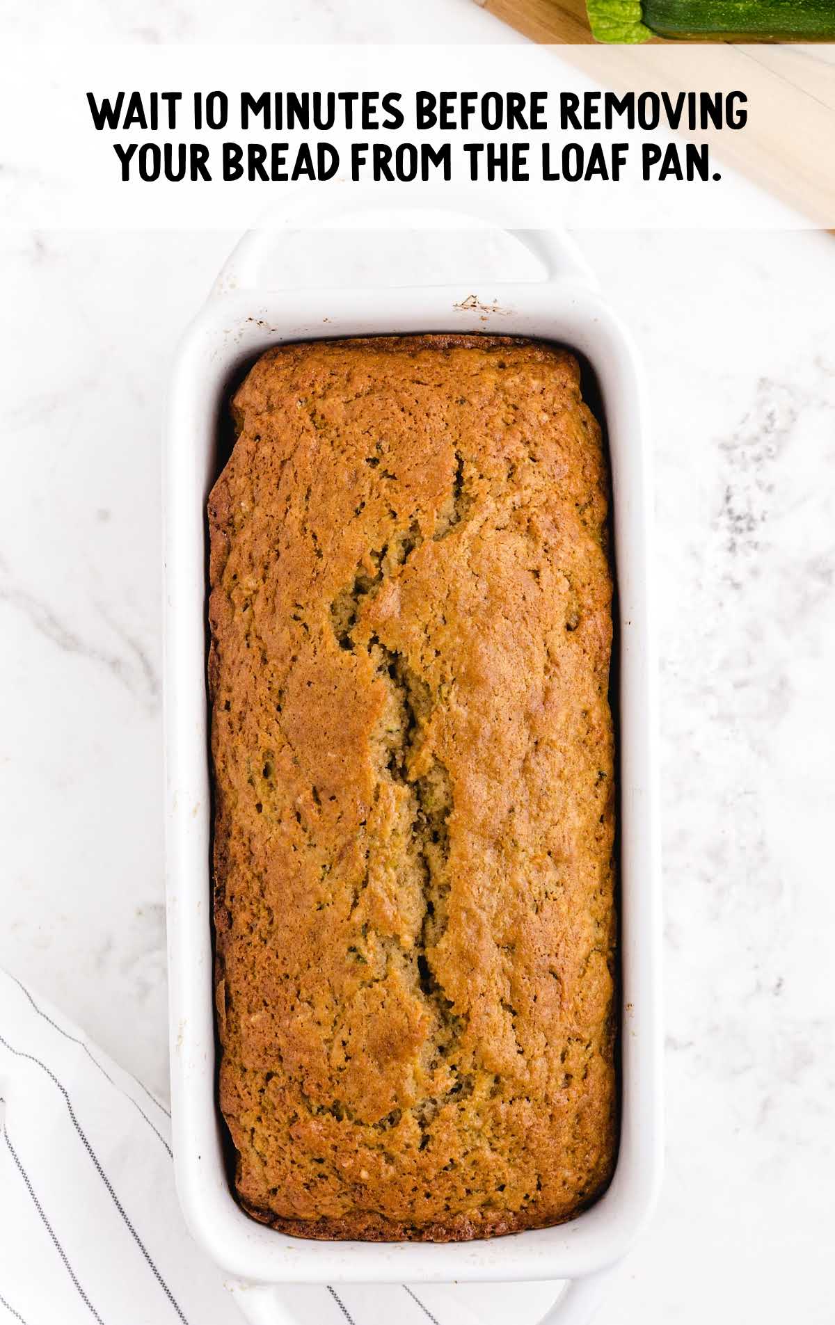 Loaf of freshly baked zucchini bread in a white pan on a marble surface with text overlay instructing to wait 10 minutes before removing from the pan.
