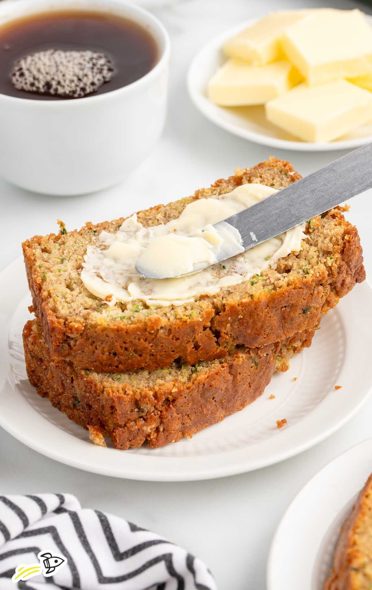 Zucchini bread slices stacked on a plate with one slice being spread with butter and a cup of tea in the background.