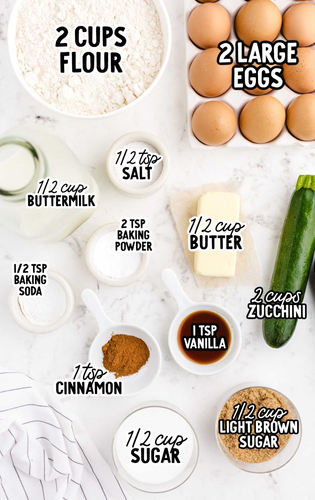 Overhead shot of zucchini bread ingredients including flour, eggs, zucchini, butter, sugars, and spices labeled on a marble surface.
