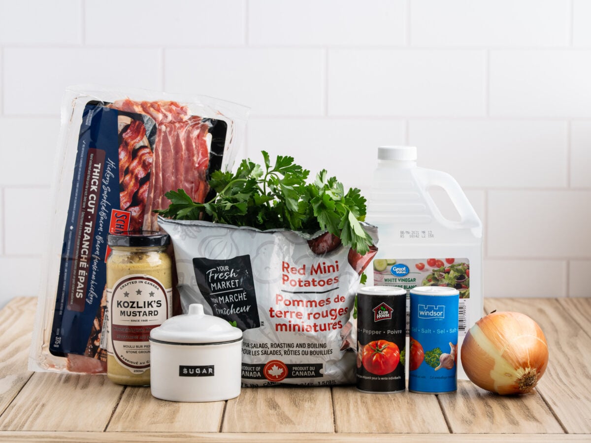 A variety of ingredients for a German Potato Salad Recipe on a wooden table—bacon, mini red potatoes, mustard, parsley, white vinegar, canned tomatoes, salt, sugar, pepper, and yellow onion—set against a white tiled background.