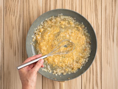 A hand holding a whisk stirs a mixture of chopped onions and creamy yellow sauce in a frying pan on a light wooden surface, just like the beginning steps of an authentic German Potato Salad Recipe.