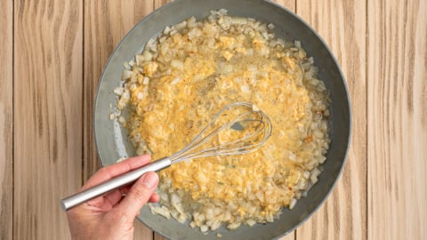 A hand holding a whisk stirs a mixture of chopped onions and creamy yellow sauce in a frying pan on a light wooden surface, just like the beginning steps of an authentic German Potato Salad Recipe.