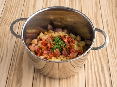 A stainless steel pot on a wooden surface contains ingredients for a classic German Potato Salad Recipe—chopped potatoes, bacon pieces, herbs, and vegetables—ready to be cooked or mixed.