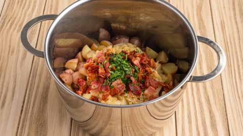 A stainless steel pot on a wooden surface contains ingredients for a classic German Potato Salad Recipe—chopped potatoes, bacon pieces, herbs, and vegetables—ready to be cooked or mixed.
