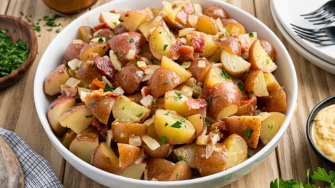 A white bowl filled with chopped German Potato Salad, garnished with parsley and bits of bacon, sits on a wooden table surrounded by plates, forks, fresh herbs, and small bowls of condiments.
