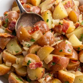 A bowl of German Potato Salad Recipe features sliced red potatoes, crispy bacon pieces, chopped onions, and fresh herbs, with a large spoon scooping up a hearty portion.
